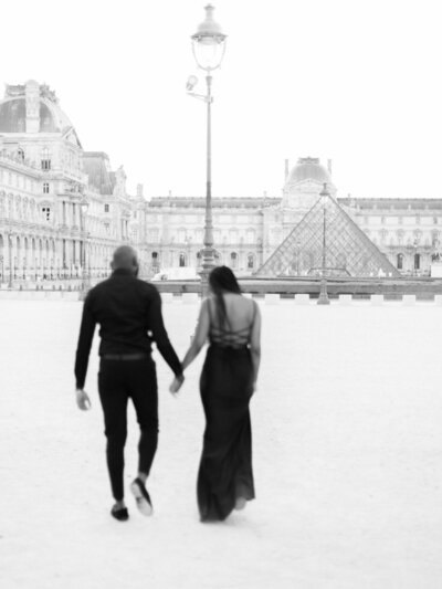 Un couple traverse la cour du Louvre, robe longue noire et costume sombre.
